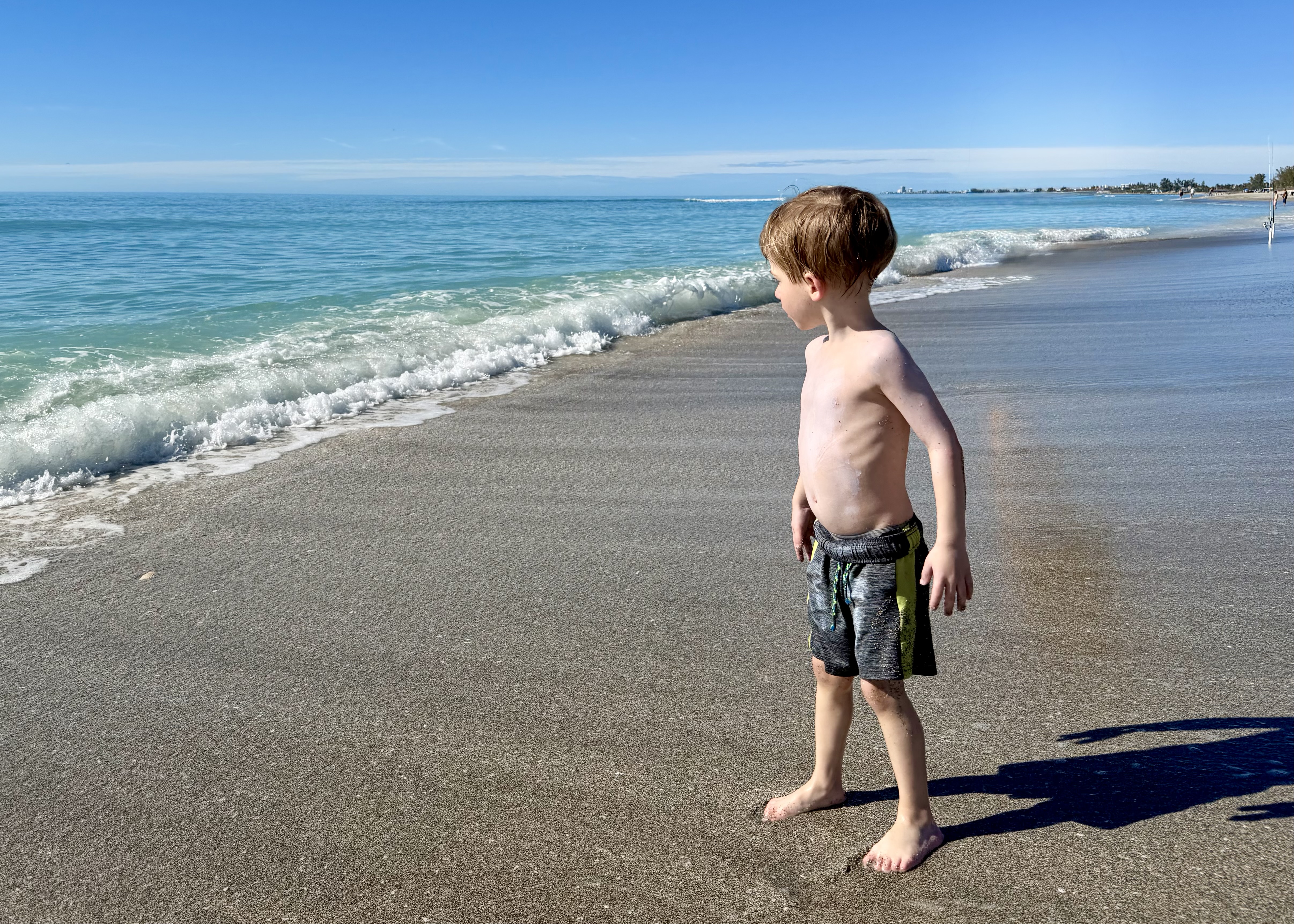 A young boy in swim trunks stands barefoot at the shoreline of a clear turquoise beach