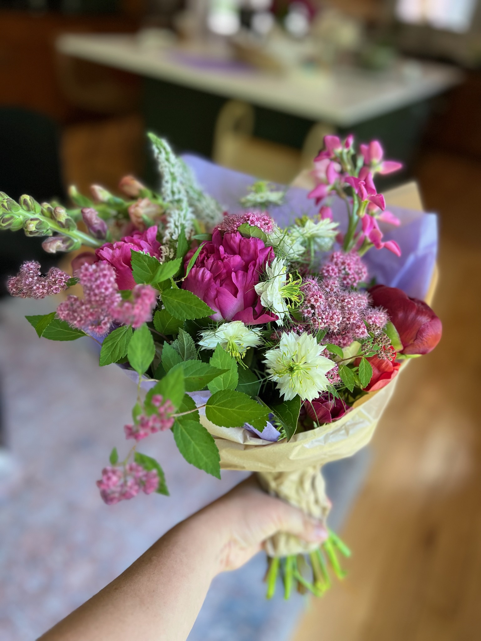 A hand holds a lush bouquet of magenta peonies, white nigella, and snapdragons wrapped in kraft paper