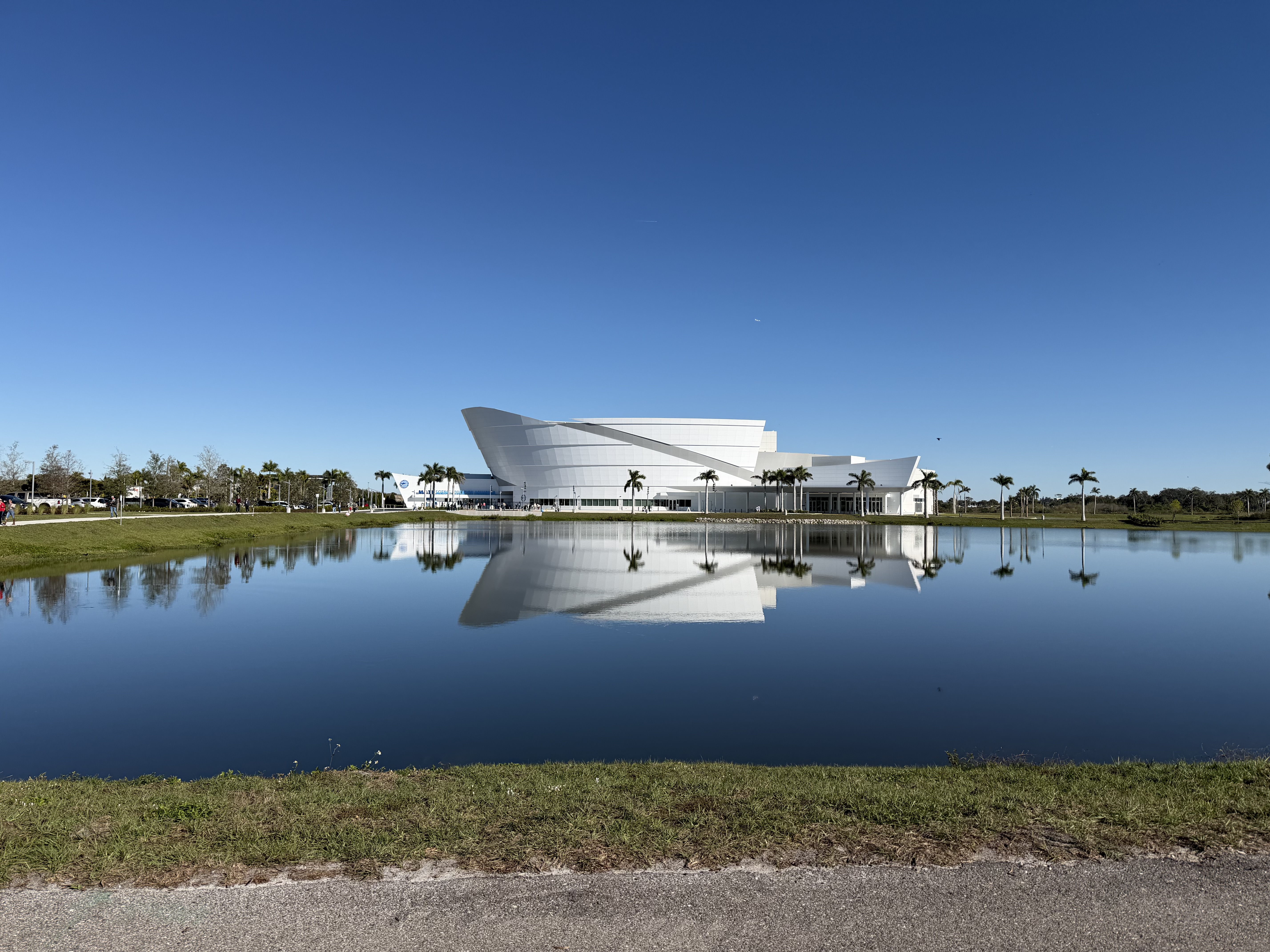 A sleek, modern white arena reflects perfectly in a still pond under a clear blue Florida sky surrounded by palm trees
